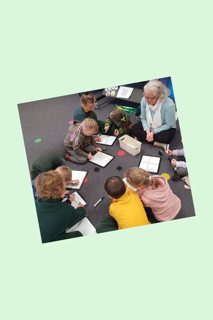 A teacher sitting with a group of students working on whiteboard for a Kinder Start day with older and younger children.