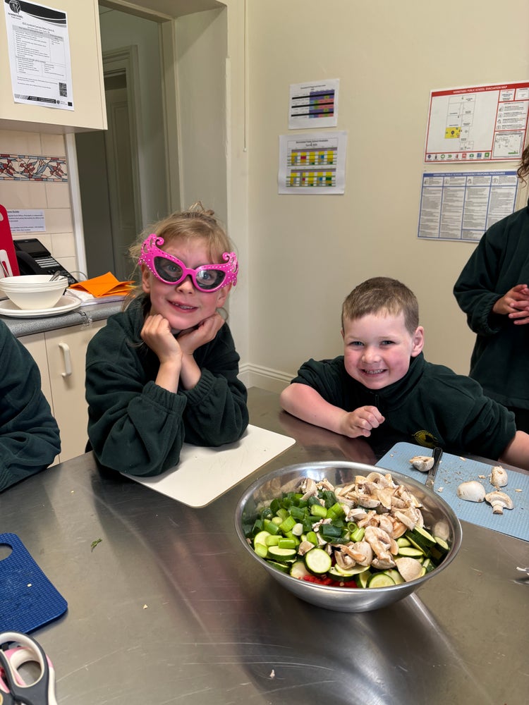 Two smiling students with a bowl of chopped up vegetables in front of them. One student is wearing special glasses as she was cutting onions.